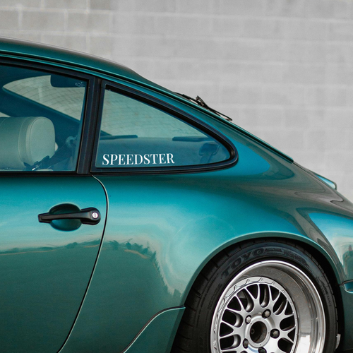 Teal car with 'Speedster' window sticker parked against a gray wall.